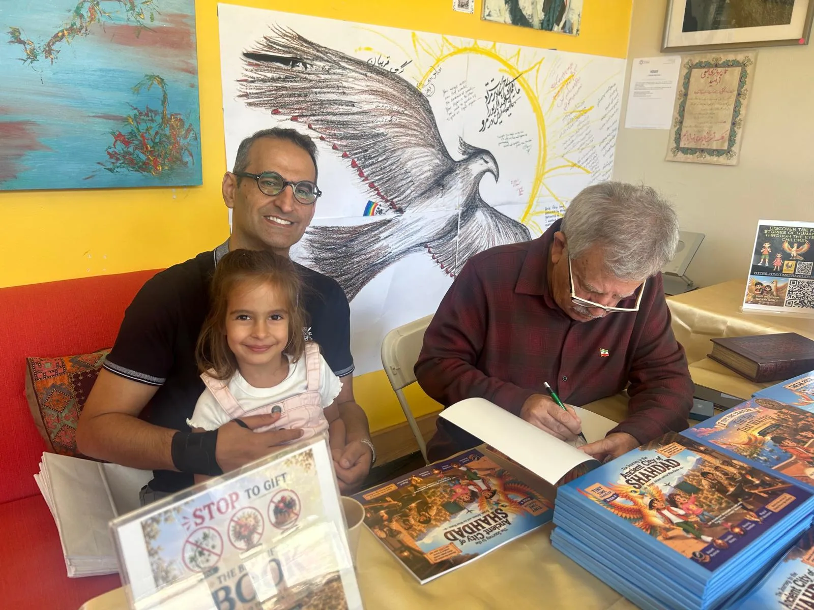 Dr. Bahram Ghadiri signs a copy of The Journey to the Ancient City of Shahdad at the Stories, Songs & Imagination event in Berkeley, with a smiling young reader and her family beside him and a large Simurgh drawing covered in handwritten notes on the wall behind.